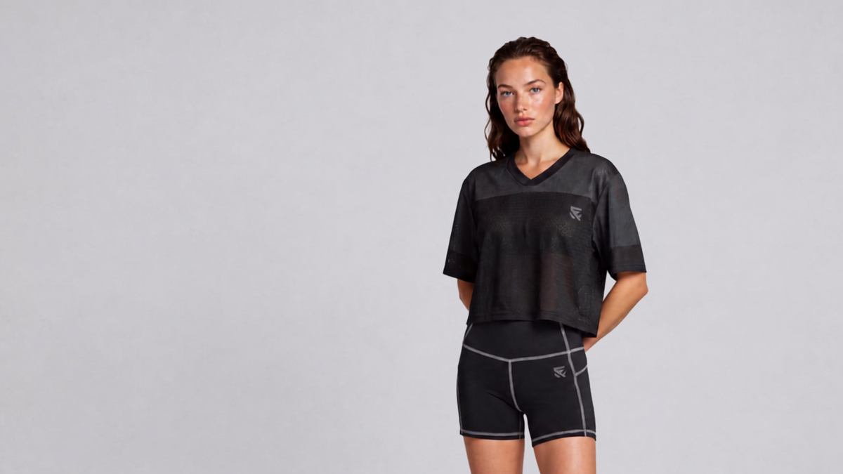 A woman modeling a black mesh cropped V-neck athletic top and matching black athletic shorts with light gray piping, photographed against a plain light gray studio background.