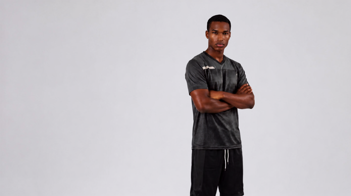 A man stands against a light gray studio background with his arms crossed, wearing a dark gray athletic t-shirt and black athletic pants.